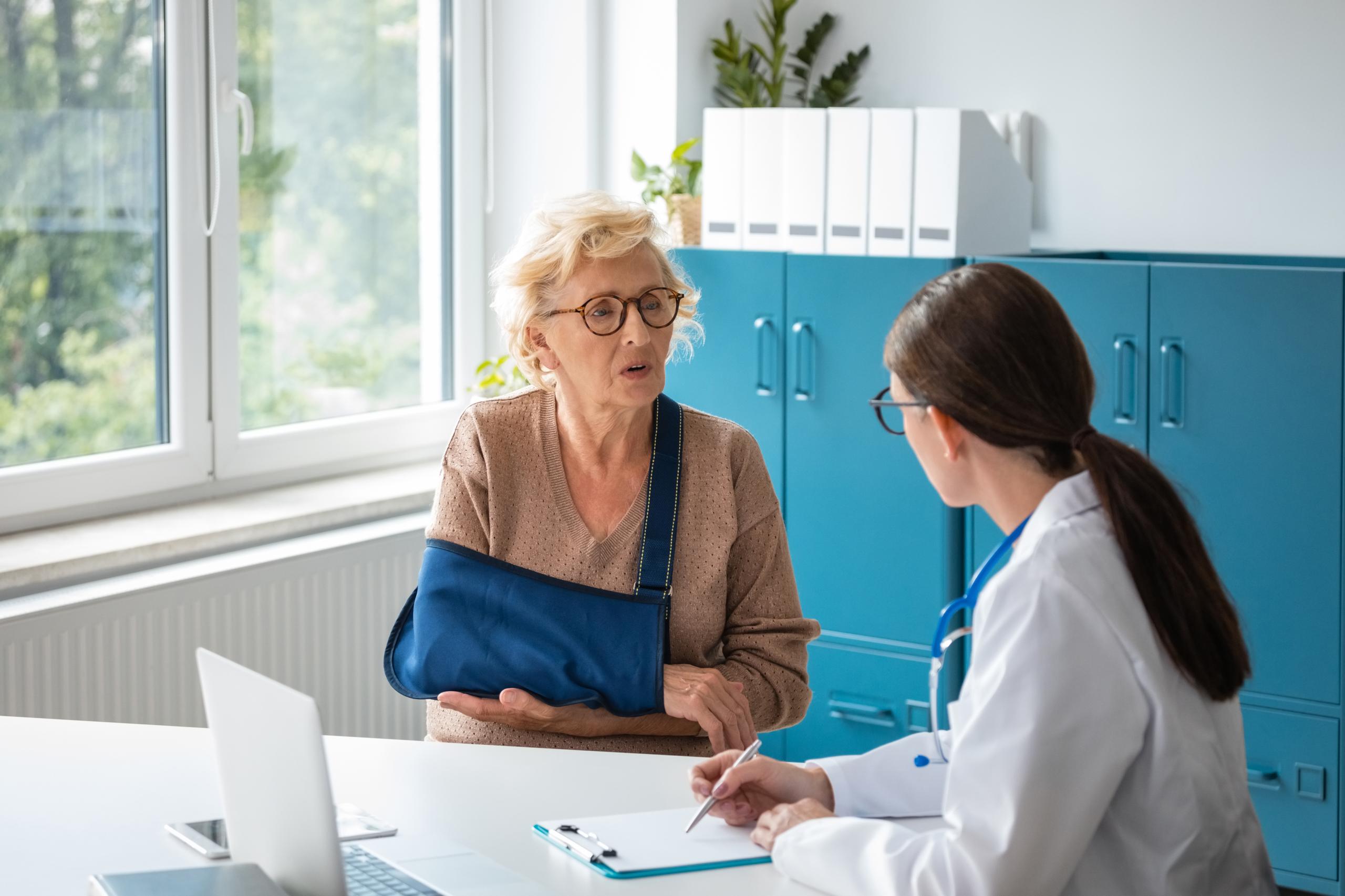 A woman with an arm in a cast, sitting at a doctors desk and speaking to the doctor